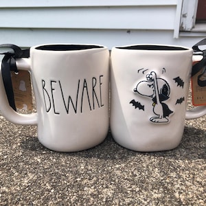 May include: Two white ceramic mugs with black interiors. One mug says "BEWARE" in black letters. The other mug features a Snoopy design in a Halloween costume with bats. Both mugs have black bows and tags attached.