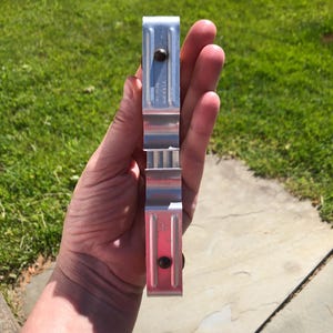 May include: A silver-colored metal bracket with a rectangular, channel-like profile. The piece features two dark, circular fasteners positioned along its length and embossed text that reads "PAT 37744" and "MADE IN USA".