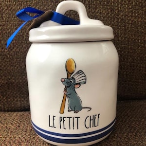 May include: White ceramic canister with a blue and white striped lid and base. The canister features a cartoon illustration of Remy the rat from the movie Ratatouille, holding a wooden spoon and wearing a chef's hat. The text "Le Petit Chef" is printed on the front of the canister.