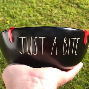 Puede incluir: Un cuenco de cerámica negra con el texto "JUST A BITE" escrito en letras blancas. El cuenco presenta dos detalles rojos brillantes en forma de goteo en el borde superior, que recuerdan a la sangre, situados a ambos lados del exterior.