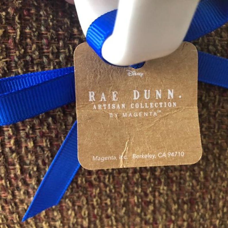 May include: A brown paper tag with the text "RAE DUNN. ARTISAN COLLECTION BY MAGENTA" and "Magenta, Inc. Berkeley, CA 94710". The tag is tied with a blue ribbon.