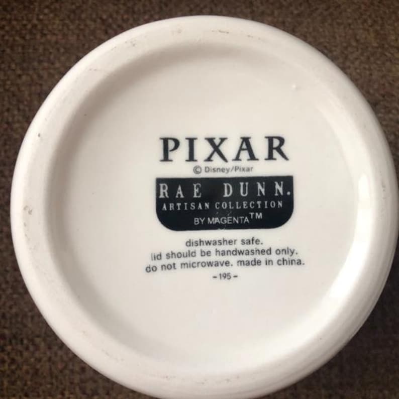 May include: White ceramic mug with a black Pixar logo, Rae Dunn. Artisan Collection by MagentaTM. Dishwasher safe, handwash lid only, do not microwave, made in China. -195-
