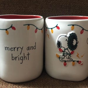 May include: Two white ceramic mugs with red interiors and handles. One mug has the text "merry and bright", the other features Snoopy tangled in Christmas lights. Both mugs have a string of Christmas lights around the rim.