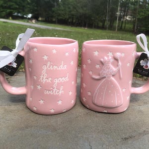 May include: Two light pink ceramic mugs featuring white star patterns. One mug displays the embossed text 'glinda the good witch' in white, while the other features an embossed white figure of a witch holding a wand. Both mugs have white ribbons.