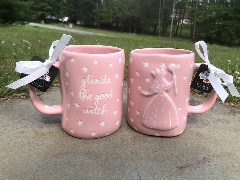 May include: Two light pink ceramic mugs featuring white star patterns. One mug displays the embossed text 'glinda the good witch' in white, while the other features an embossed white figure of a witch holding a wand. Both mugs have white ribbons.