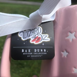 May include: A light pink ceramic mug featuring embossed white stars and a white ribbon tied around the handle. A black product tag is attached, displaying the text The Wizard of Oz with red slippers, followed by Rae Dunn Artisan Collection by Magenta.