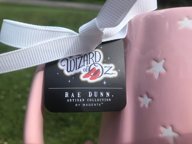 May include: A light pink ceramic mug featuring embossed white stars and a white ribbon tied around the handle. A black product tag is attached, displaying the text The Wizard of Oz with red slippers, followed by Rae Dunn Artisan Collection by Magenta.