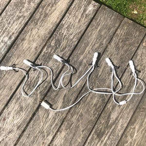 May include: A string of white decorative lights featuring six individual bulb sockets connected by a white electrical cord. The set includes a standard two-prong electrical plug at one end, designed for indoor or outdoor illumination purposes.