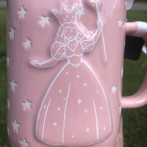 May include: A light pink ceramic mug featuring a white embossed design of a figure wearing a crown and holding a star-tipped wand. The surface is decorated with numerous small, recessed white stars scattered across the exterior of the mug.