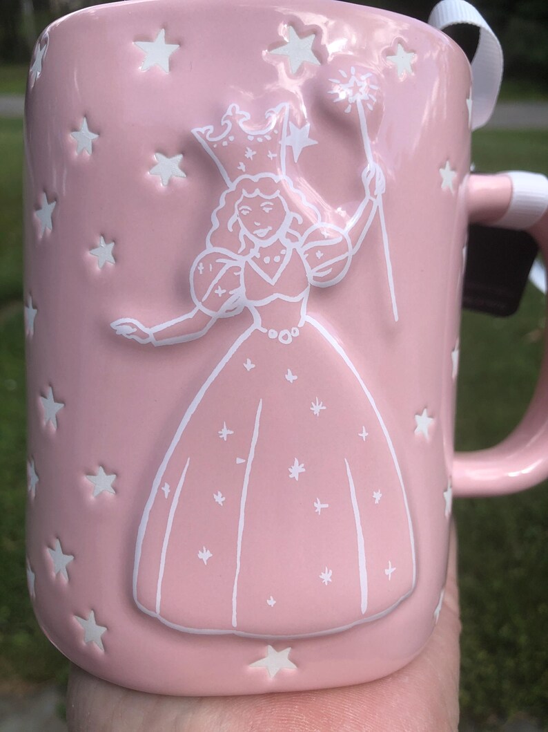 May include: A light pink ceramic mug featuring a white embossed design of a figure wearing a crown and holding a star-tipped wand. The surface is decorated with numerous small, recessed white stars scattered across the exterior of the mug.