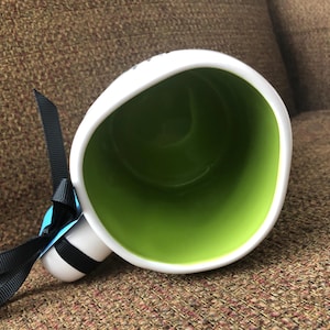 Puede incluir: Taza de cer&aacute;mica blanca con asa de cinta negra y un interior verde. La taza est&aacute; sobre una superficie de tela marr&oacute;n.