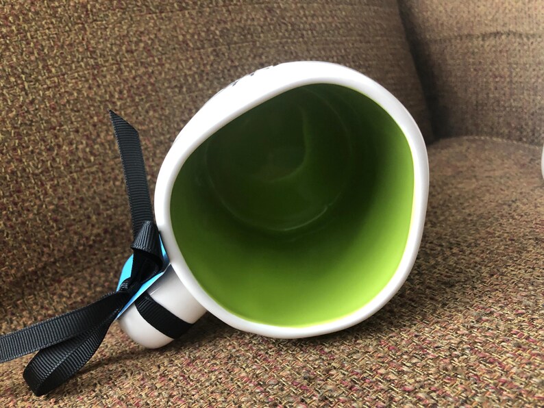 Puede incluir: Taza de cer&aacute;mica blanca con asa de cinta negra y un interior verde. La taza est&aacute; sobre una superficie de tela marr&oacute;n.