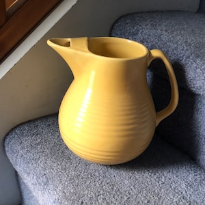 May include: A yellow ceramic jug with a rounded body and a wide spout. The jug has a smooth, glossy finish and a slightly textured surface.