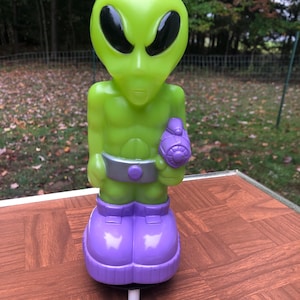 Puede incluir: Un juguete de extraterrestre verde con un traje espacial morado y un cinturón plateado. El extraterrestre sostiene una pistola espacial plateada y negra. El juguete está sobre una superficie a cuadros marrón y blanca.