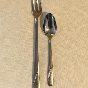 May include: A stainless steel fork and spoon set. The fork has three tines, and the spoon has a rounded bowl. Both utensils have a simple, elegant design with a curved accent on the handle. The set is placed on a neutral-colored surface.