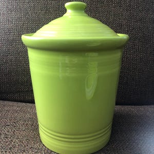 Puede incluir: Un bote de cerámica verde brillante con tapa a juego. El bote tiene forma cilíndrica con un borde ligeramente ensanchado y una serie de estrías horizontales cerca de la base. La tapa tiene un pequeño pomo.