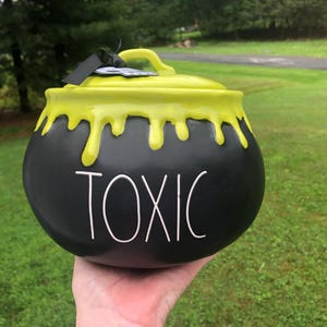 Pode incluir: Um recipiente de cerâmica preto em forma de caldeirão com tampa, apresentando um padrão de gotejamento verde-limão brilhante na borda superior. A palavra TOXIC está impressa em letras maiúsculas brancas, altas e finas, na parte frontal.