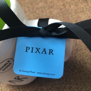 Puede incluir: Una etiqueta azul con la palabra "PIXAR" en blanco. Debajo de la palabra est&aacute; el texto "&copy; Disney/Pixar www.disney.com". La etiqueta est&aacute; atada a una taza blanca con una cinta negra.