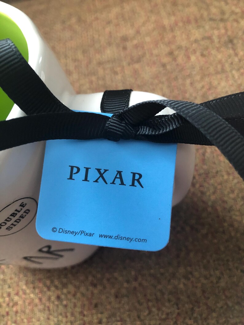 Puede incluir: Una etiqueta azul con la palabra "PIXAR" en blanco. Debajo de la palabra est&aacute; el texto "&copy; Disney/Pixar www.disney.com". La etiqueta est&aacute; atada a una taza blanca con una cinta negra.