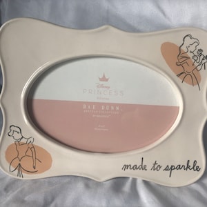 May include: Cream-colored ceramic photo frame with an oval opening. Features Disney Princess branding and illustrations of princesses. The frame has a decorative edge and the words "made to sparkle" are written on the bottom.