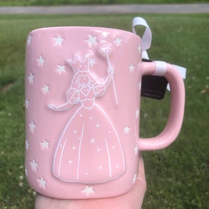 May include: A light pink ceramic mug featuring a raised white embossed design of a fairy wearing a crown and holding a star-tipped wand. The mug is decorated with numerous small white embossed stars scattered across the surface and includes a white ribbon tied to the handle.
