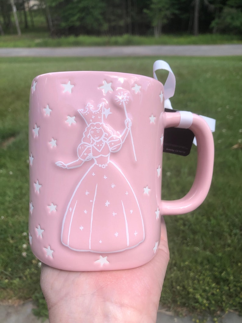 May include: A light pink ceramic mug featuring a raised white embossed design of a fairy wearing a crown and holding a star-tipped wand. The mug is decorated with numerous small white embossed stars scattered across the surface and includes a white ribbon tied to the handle.