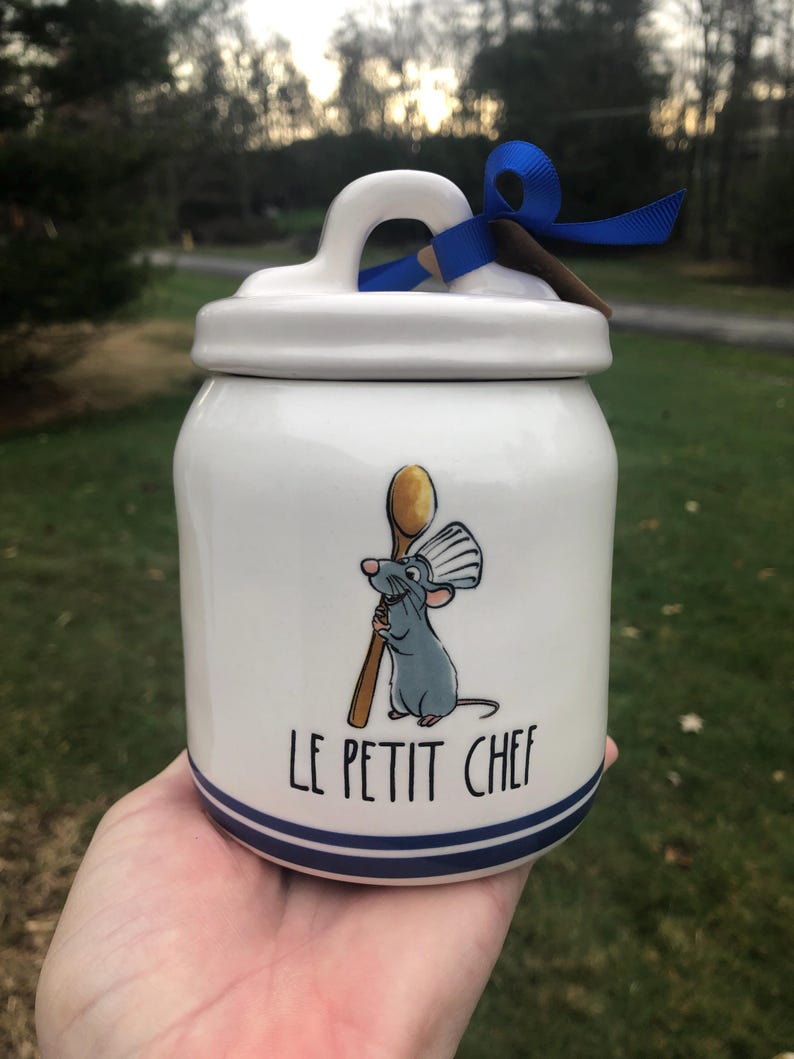 May include: White ceramic jar with a blue and white striped lid and a blue ribbon. The jar features a cartoon rat holding a wooden spoon and the text "Le Petit Chef".