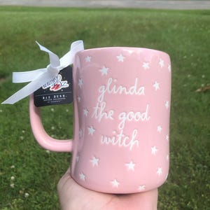 May include: A light pink ceramic mug featuring white embossed stars and the text "glinda the good witch" in a cursive font. A white ribbon is tied to the handle, attached to a black tag with the "Wizard of Oz" logo and "Rae Dunn" branding.