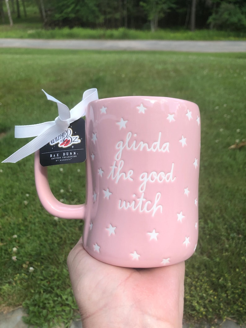May include: A light pink ceramic mug featuring white embossed stars and the text "glinda the good witch" in a cursive font. A white ribbon is tied to the handle, attached to a black tag with the "Wizard of Oz" logo and "Rae Dunn" branding.