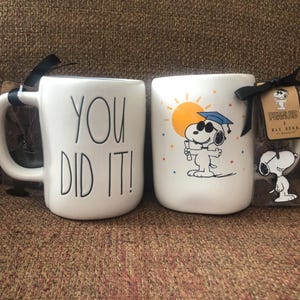 Pode incluir: Duas canecas de cerâmica brancas. Uma caneca tem o texto "YOU DID IT!" em preto. A outra caneca apresenta Snoopy com um chapéu de formatura e um diploma, e o texto "PEANUTS x Rae Dunn". Uma pequena figura de Snoopy está anexada.