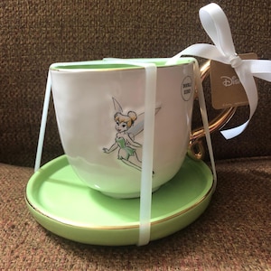 May include: A white teacup and saucer set featuring a gold handle and rim, and a green saucer. The teacup has a cartoon image of Tinkerbell. The set is tied with a white ribbon and a Disney tag.