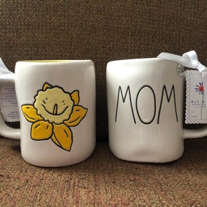 May include: Two white ceramic mugs. One mug features a yellow daffodil design with a smiling face. The other mug has the word "MOM" in black lettering. Both mugs have a white ribbon tied around the handle.