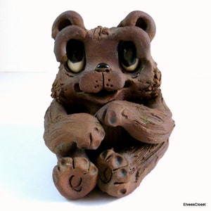 May include: A brown ceramic bear figurine with large eyes and a wide smile. The bear is sitting with its paws crossed in front of it.