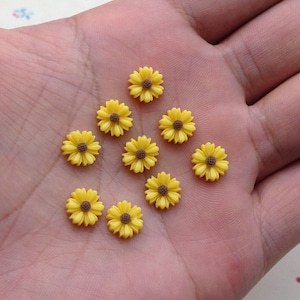 May include: A collection of ten small yellow resin sunflower embellishments with brown centers. The flowers are arranged in a triangular pattern on a hand.