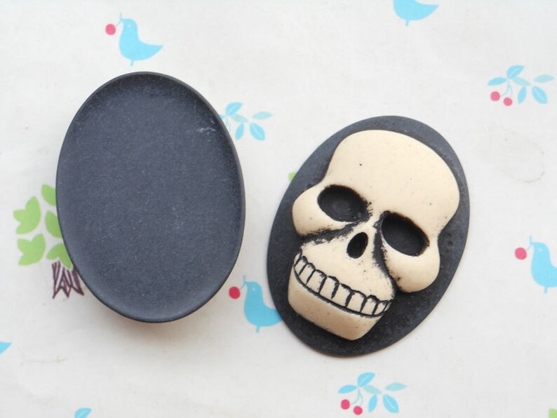 May include: Two black and white skull cabochons. One skull is on a black background and the other is on a white background.