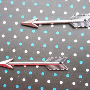 May include: Two silver arrow charms with a textured design. The arrows are on a black background with white and blue polka dots.
