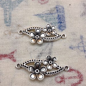 May include: Two silver-toned metal flower charms with a decorative design. Each charm features two five-petal flowers with a small circle in the center. The charms have two loops on the sides for attaching to jewelry or other crafts.
