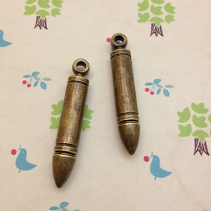 Puede incluir: Dos abalorios de color dorado antiguo en forma de bala con un bucle en la parte superior para sujetarlos a joyas u otras manualidades.