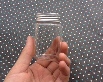 Puede incluir: Un frasco de vidrio transparente con tapa plateada. El frasco está vacío y se sostiene en una mano.
