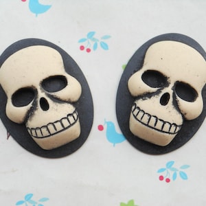 May include: Two black and white skull-shaped cabochons with a smiling expression. The skulls are set against a black background.