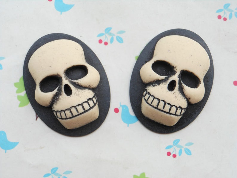 May include: Two black and white skull-shaped cabochons with a smiling expression. The skulls are set against a black background.
