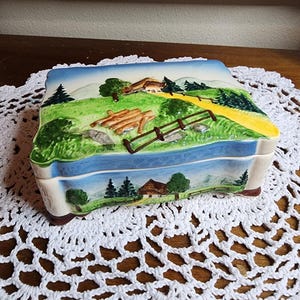 May include: A rectangular ceramic trinket box with a landscape design. The lid features a painted scene of a house, trees, a fence, and a road. The box is primarily white, green, blue, and brown.
