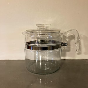 May include: A clear glass coffee pot with a silver band around the middle. The pot has a glass lid and a glass handle.