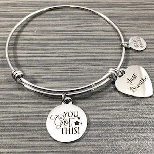May include: Silver expandable bangle bracelet with three charms. The charms are a heart that says "Just Breathe", a round charm that says "You Got This!" with stars, and a smaller round charm that says "Keep" on one side and "Smile" on the other.