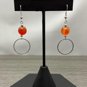 May include: A pair of dangle earrings featuring a faceted orange gemstone bead above a silver-tone metal hoop. The earrings hang from silver-tone ear wires. The earrings are displayed on a black stand against a neutral background.