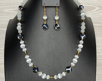 May include: A necklace and earring set displayed on a black mannequin. The necklace features white and black patterned beads, separated by gold-colored bars. The earrings have a similar design, with white and black beads suspended from gold-colored hooks.