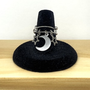May include: A silver ring with a crescent moon charm and three star charms dangling from it. The ring is displayed on a black velvet ring holder.