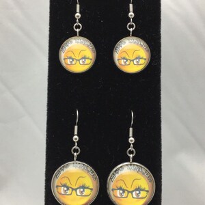 May include: A pair of silver dangle earrings with yellow round glass charms. The charms feature a cartoon face with glasses and the text "Eye Roll" written around the face.