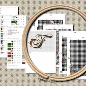 Può includere: Un telaio da ricamo in legno posizionato sopra una raccolta di fogli con schemi a punto croce stampati. I fogli mostrano una lettera T decorativa con accenti floreali, grafici a griglia in bianco e nero e una legenda dei colori.