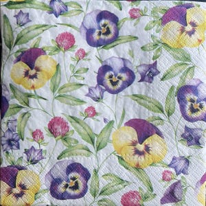 Puede incluir: Servilleta floral blanca con un estampado de pensamientos morados y amarillos, tréboles rosas y hojas verdes. Los pensamientos tienen centros morados oscuros y la servilleta tiene una superficie texturizada.